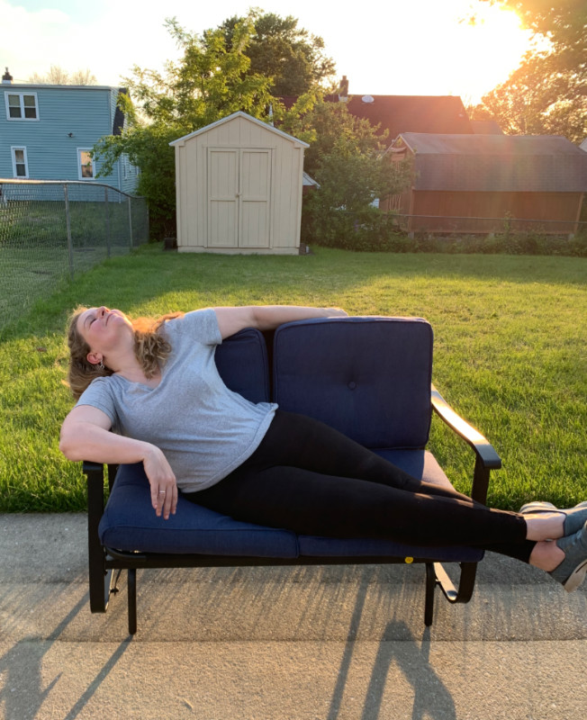 Kim, in a tee and leggings, lounging dramatically across a 2-seat glider in a back yard. It's the golden hour and the sun is streaming across the grass.