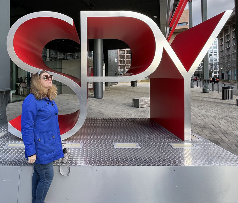 Kim posing in front of a large statue that says SPY. She is wearing a bright blue coat and gaudy sunglasses.