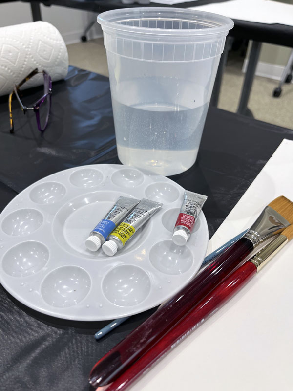 watercolor class supplies including a soup container full of water, a plastic palette from Michael's that's worthless, three tiny tubes of pigment and some low-grade brushes