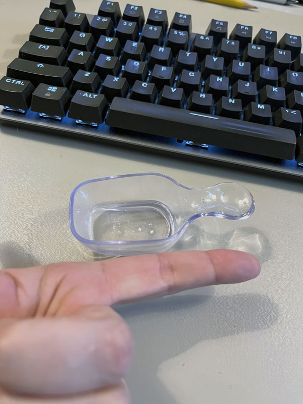 a clear plastic scoop with my index finger next to it for scale. It's smaller than my finger