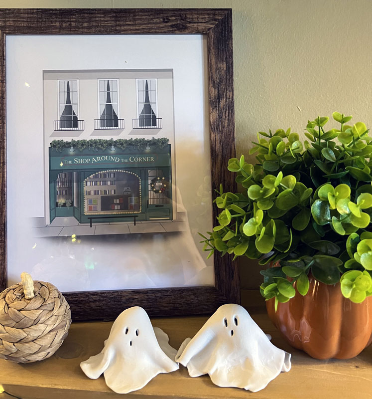 two clay ghosts on a shelf with tiny wicker pumpkins, a fake plant in a pumpkin flowerpot, and a "the shop around the corner" print - an homage to You've Got Mail.
