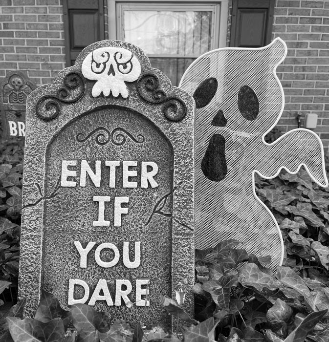 a black and white photo of Halloween decorations. The foam gravestone says "ENTER IF YOU DARE" and there is a metal ghost peeking out from behind it. They are both placed into the ivy patch outside of my house.