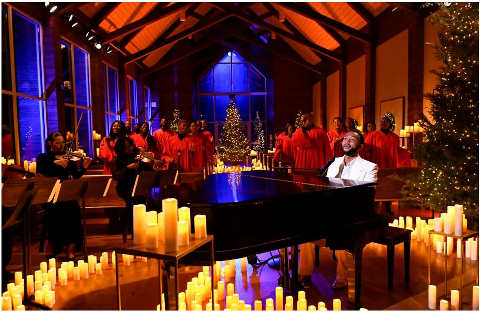 John Legend at a piano in Elvis's chapel. He's in white, piano is black, dozens of white candles surround him. There's a choir spaced through out the pews behind him. They are in red. At the center in the background is a Christmas tree.
