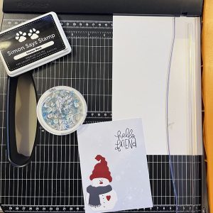 a paper cutter, stamp pad, snowflake sequins, and a finished card with a snowman on it