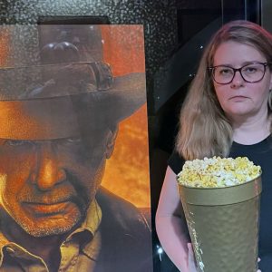 Kim is grumpily standing next to a poster of Indiana Jones and the DIial Destiny. She is holding an expensive souvenir popcorn bucket.