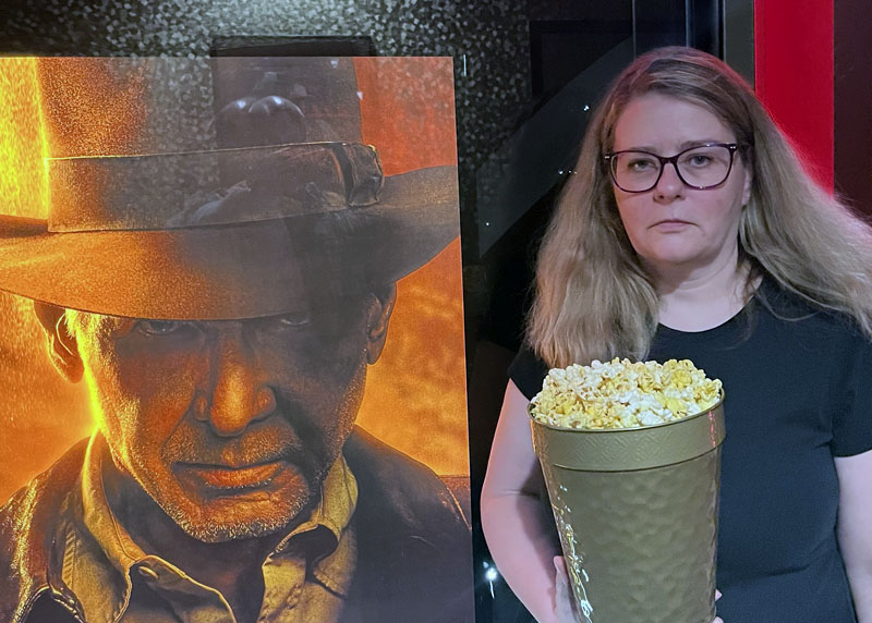 Kim, looking miserable next to a poster of Indiana Jones and the Dial of Destiny. She is holding a souvenir popcorn bucket.