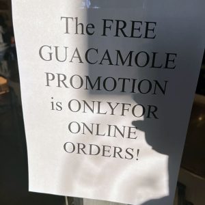 a hastily made flyer taped to the door of Chipotle that says "THE FREE GUACAMOLE PROMOTION is ONLYFOR ONLINE ORDERS!'
