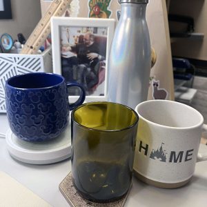 A blue mickey mouse mug, a brown glass, a natural colored mug with "HOME" on it (the o in home is a silhouette of Cinderella castle) and a S'well water bottle.