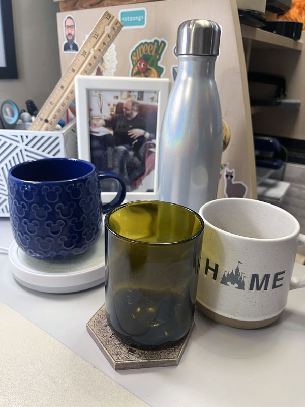 A blue mickey mouse mug, a brown glass, a natural colored mug with "HOME" on it (the o in home is a silhouette of Cinderella castle) and a S'well water bottle.