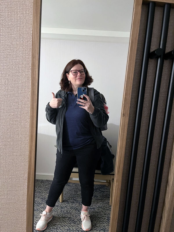 A mirror selfie of Kim giving a thumbs-up in a blue staff shirt, a gray zip up fleece, black pants and white sneakers. It is morning. 