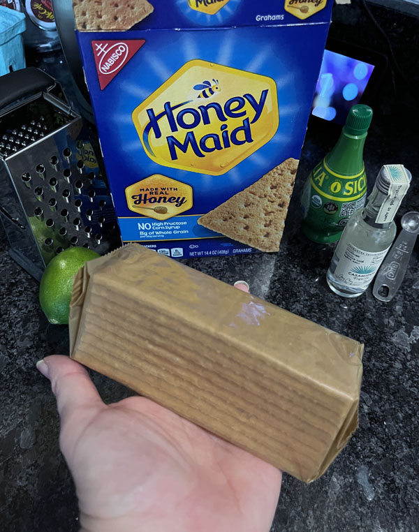 Kim holding a sleeve of Honey Maid graham crackers. Behind her hand is the box of graham crackers, a lime, a grater, a bottle of lime juice and a tiny bottle of tequila