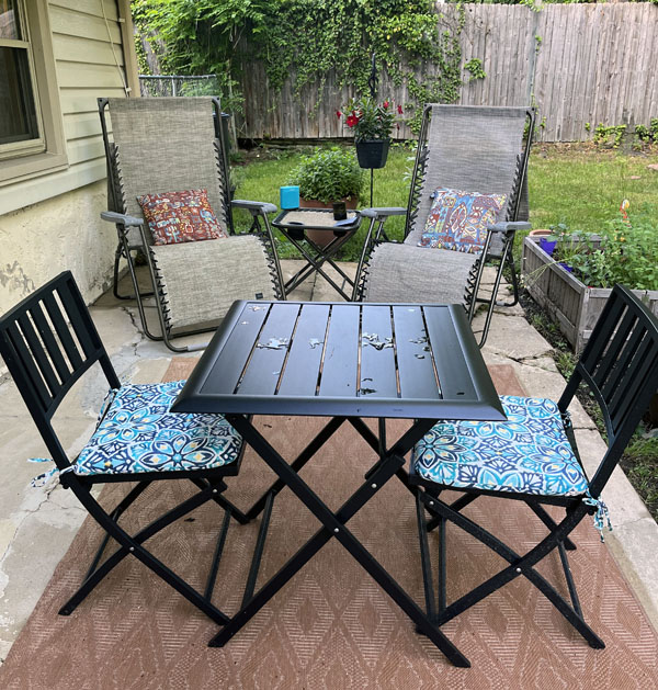My normal-shabby back patio slab, but now it has a terracotta outdoor rug, a black table & chair set with happy turquoise cushions, and two loungers with tiki pillows. There is also a hanging plant with red flowers.