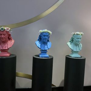 three busts of ben franklin, one in pink, one in blue, one in green. they are wearing flower headdresses