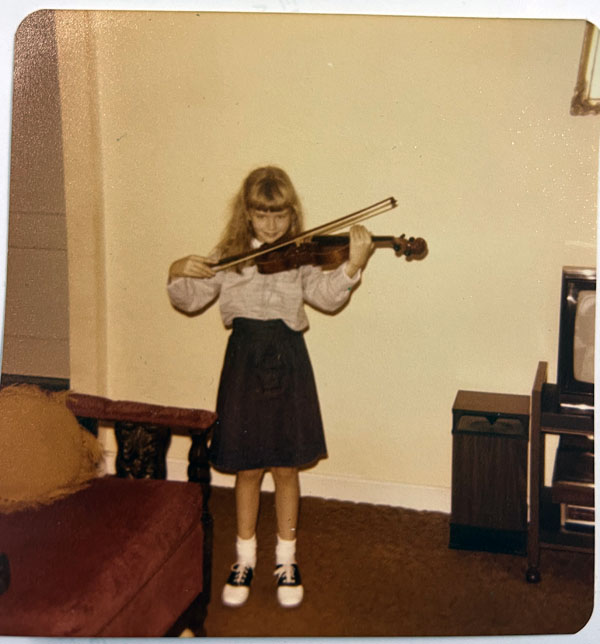 Ten year old Kim with a violin. she is wearing a blouse and knee length skirt, ankle socks and for some reason saddle shoes. a very 1970s-era red sofa is to her left, and to the right is a giant stereo speaker.