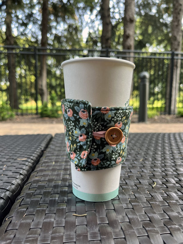 a cup of coffee with a fabric cup sleeve, sitting on an outdoor table with a woodsy tree line behind it