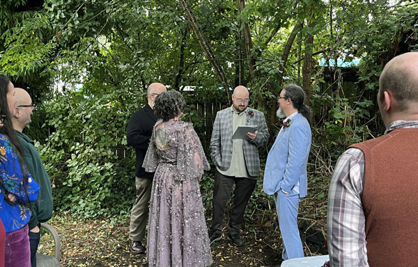 a lovely lady in a beautiful sequined dress and a lovely gentleman in a light blue suit reaffirming their love under a canopy of wet trees in front of friends