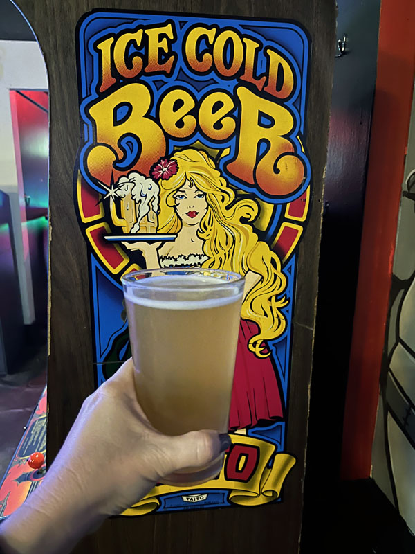 Kim holding a glass of beer in front of an "Ice Cold Beer" arcade game.