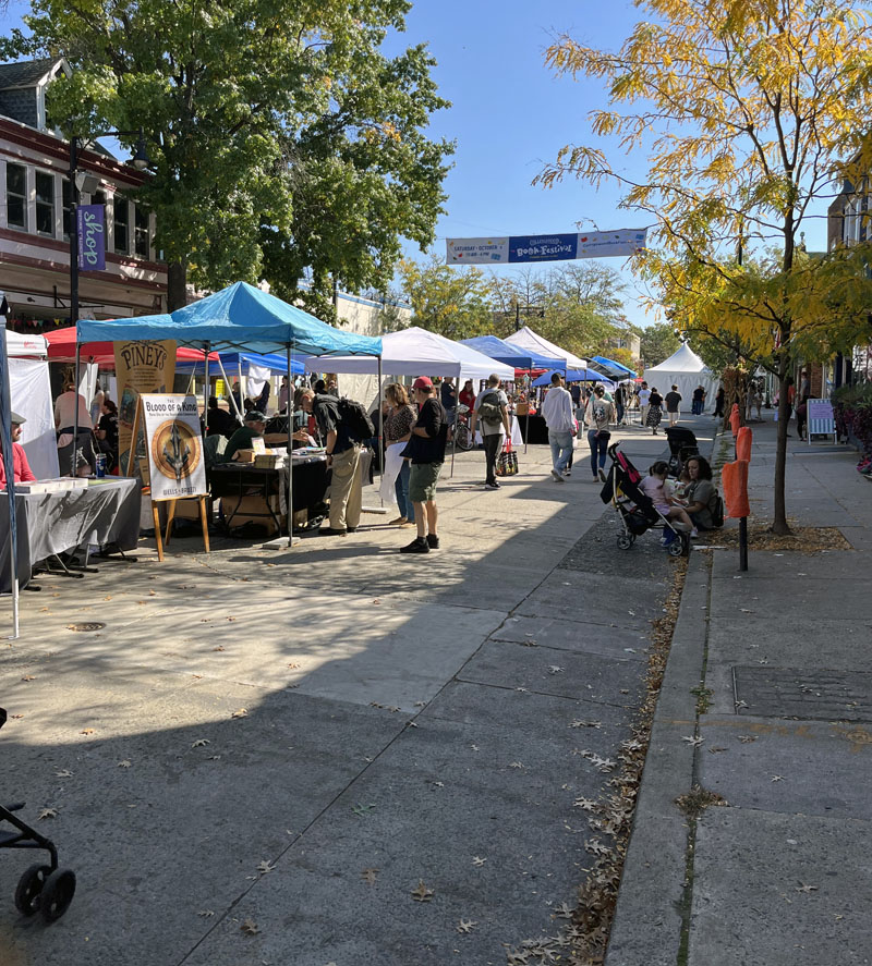 a street filled with tents and pedestrians, the banner overhead says Collingswood Book Fair. The sky is deliciously blue.