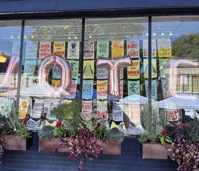 a storefront with helium balloons in the shape of letters that spell VOTE. There are many progressive posters in the window behind the balloons.