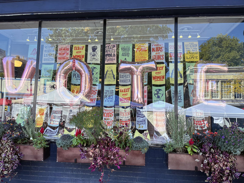 a storefront with helium balloons in the shape of letters that spell VOTE. There are many progressive posters in the window behind the balloons.