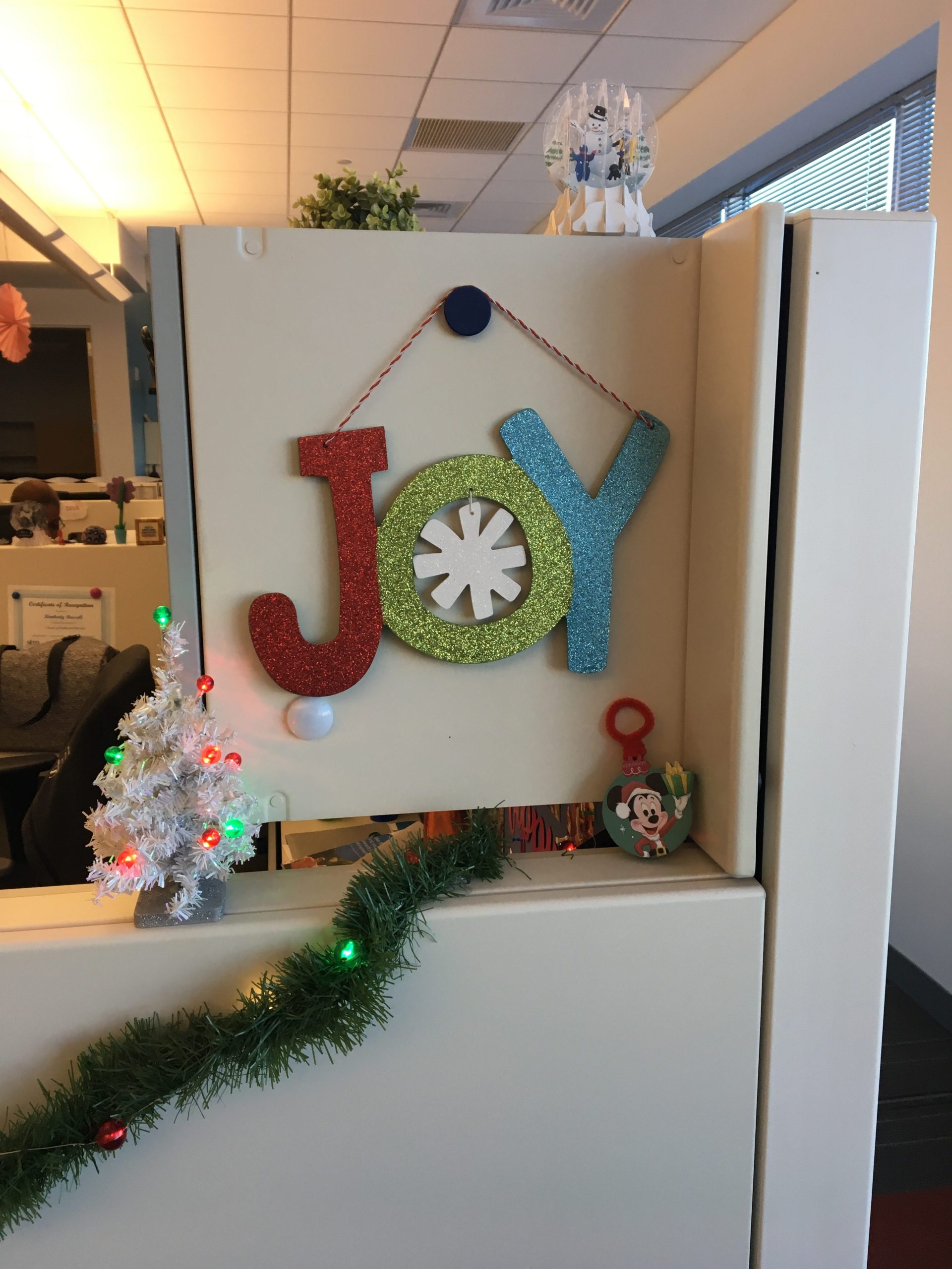 The front of my cubicle, decorated for Christmas. There is a sign that says "JOY", a small lit Christmas tree, a mickey ornament, a paper snowglobe, and a green lit garland.