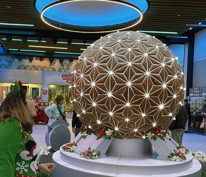 the majority of the photo is of a replica of Epcot's spaceship earth attraction in gingerbread. Kim is in a green mickey mouse christmas sweater and green minnie ears threatening to lick it.