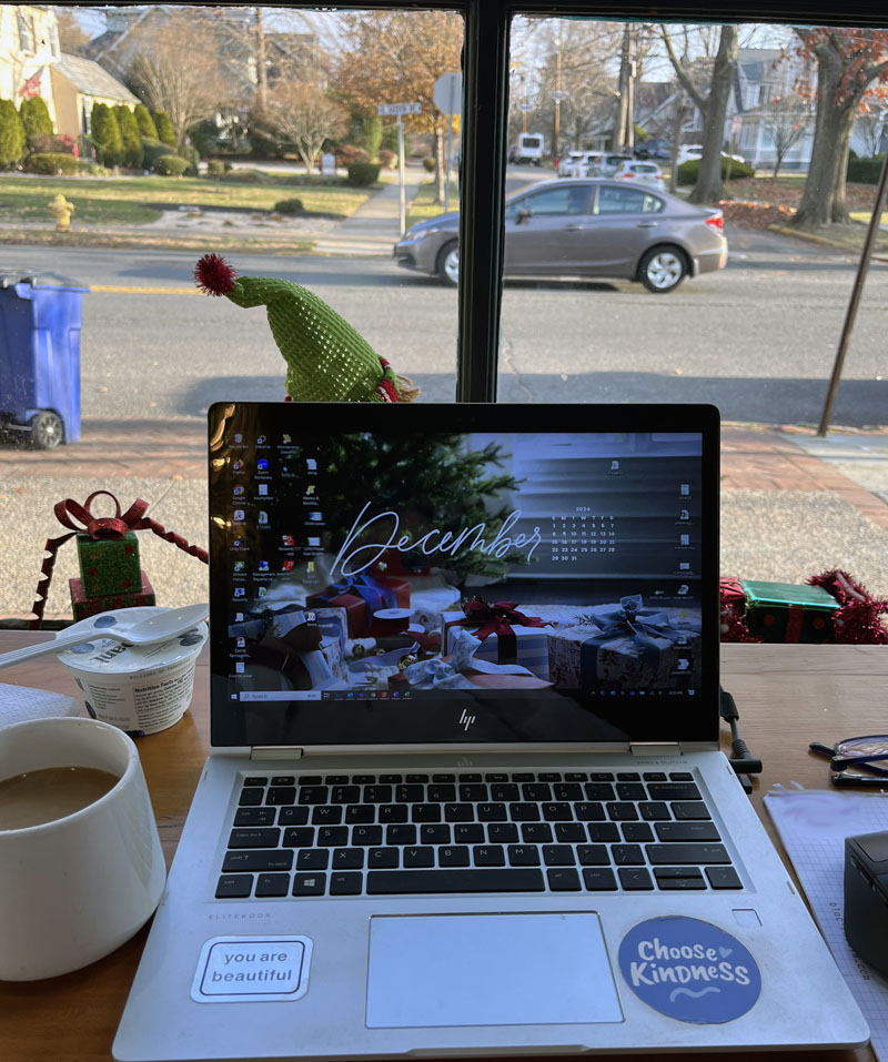 view from the coffee shop. kim's laptop is in front of her with a festive wallpaper that says december. there is an elf decoration in front of her but you only see the hat. To the left of the laptop there is a chobani yogurt and a mug of coffee. To the right there is the very edge of a notepad. behind the laptop and the elf is a large window. there is a car going by. there is a lot more natural light than the spot where kim works in her house every day, which makes her happy