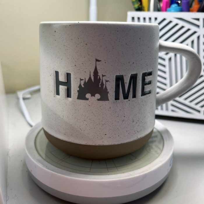 a white coffee mug with a tan bottom on top of a mug warmer. It says HOME, except the O is a silhouette of Cinderella castle at Walt Disney World.