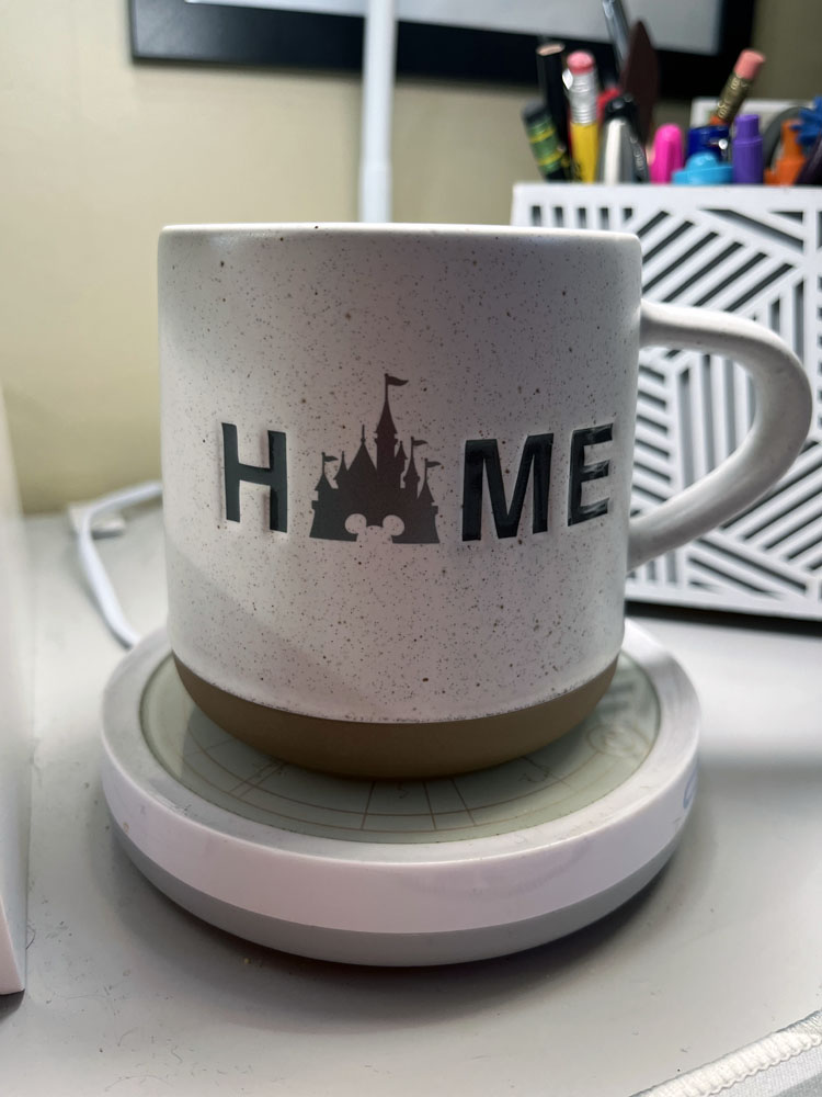 a white coffee mug with a tan bottom on top of a mug warmer. It says HOME, except the O is a silhouette of Cinderella castle at Walt Disney World.