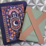 a purple kindle sleeve with pink flowers and a moon embroidered onto it. The kindle is next to it, with a sage green cover and two caramel colored silicone straps attached to the corners forming an X