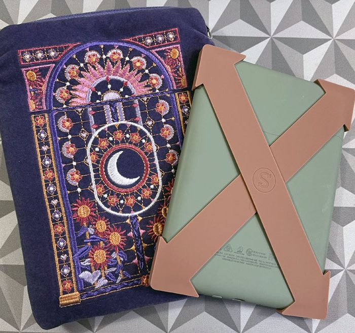 a purple kindle sleeve with pink flowers and a moon embroidered onto it. The kindle is next to it, with a sage green cover and two caramel colored silicone straps attached to the corners forming an X