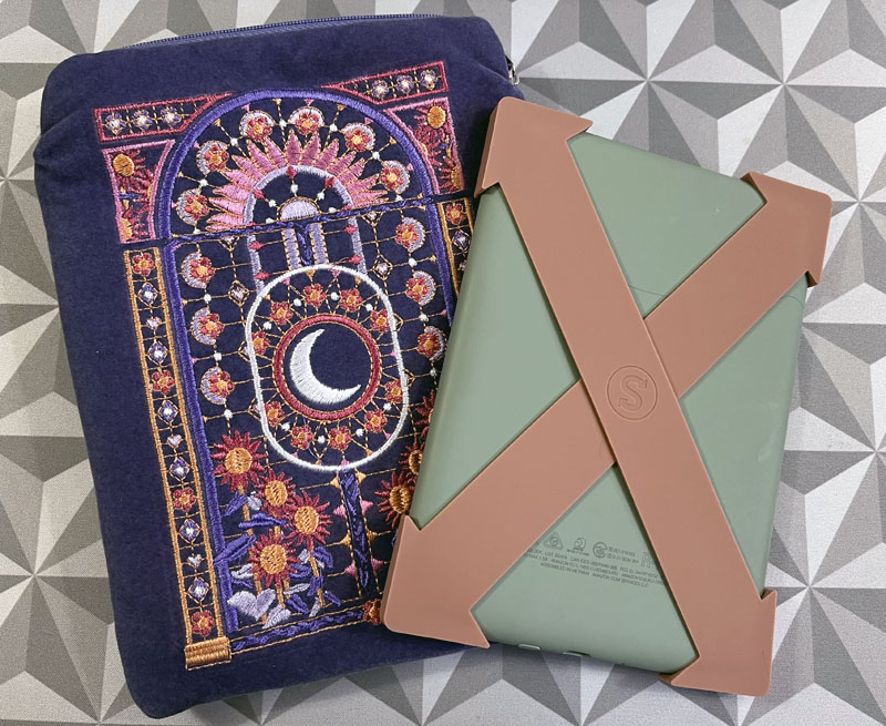 a purple kindle sleeve with pink flowers and a moon embroidered onto it. The kindle is next to it, with a sage green cover and two caramel colored silicone straps attached to the corners forming an X