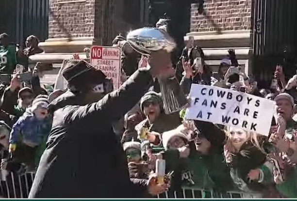 a shot from the eagles parade. A player is holding up the super bowl trophy but the real focus is a fan's sign that says "Cowboys fans are at work" Nobody likes the Cowboys.