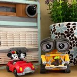 A closeup of a wood-ish shelf. There is a Lego radio, a small red car with Mickey and Minnie Mouse in it (from the runaway railway ride(), a figurine of WALL-E presenting a tiny plant in a boot, and behind him is a delicate mug with a Jonathan Adler blue and brown flower print and an artificial plant in it.