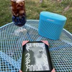 Outside. A turquoise metal side table hold a tall glass of diet dr pepper, a small turquoise Bluetooth speaker, and a Kindle displaying the cover of All Fours by Miranda July.