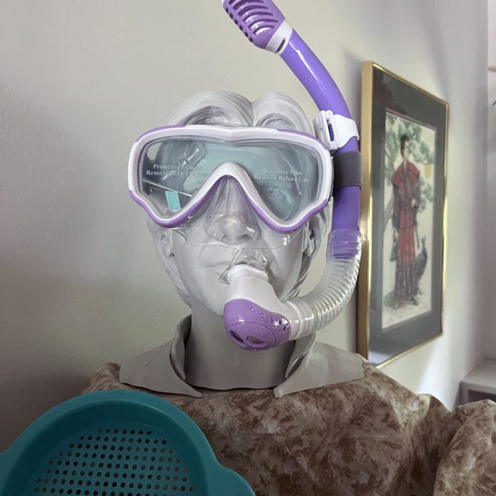 a 3d model of Harrison Ford's head, wearing a purple child's snorkel. there is a sand toy in front of it and it's on a sand-colored cloth.