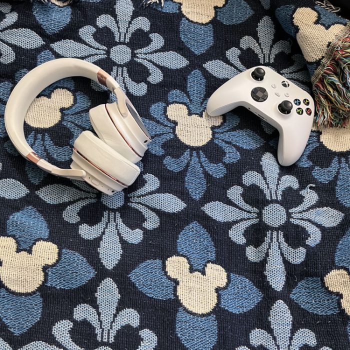 a woven throw with three shades of blue and white mickey mouse icons. On top of it are a white Xbox controller and a white pair of Raycon everyday headphones.
