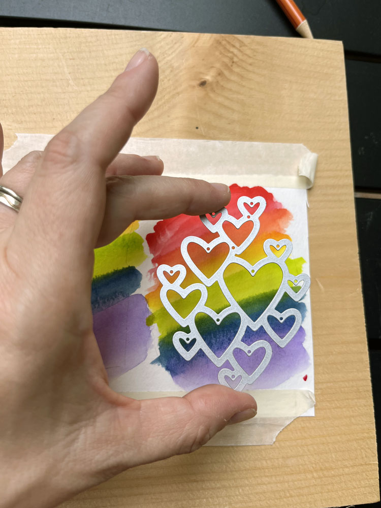 kim holding a die of a stack of hearts in front of a watercolor painted spectrum.