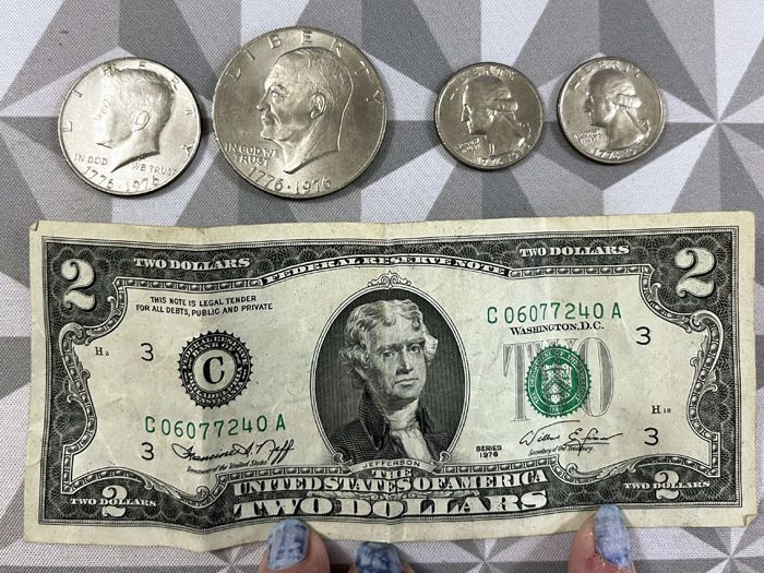 Top: four coins, a 50cent piece, a dollar coin, and two quarters. Bottom: a 2 dollar bill. All are from the 1976 bicentennial series.