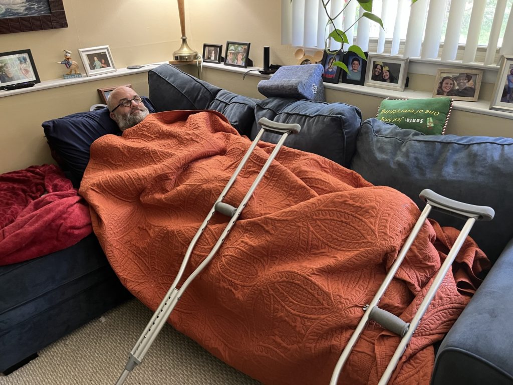 WM is lying on a blue sectional sofa. He is covered chin to toe in an orange blanket. His two crutches are propped against him to keep Murphy from leaping on him. He looks resigned to his fate.