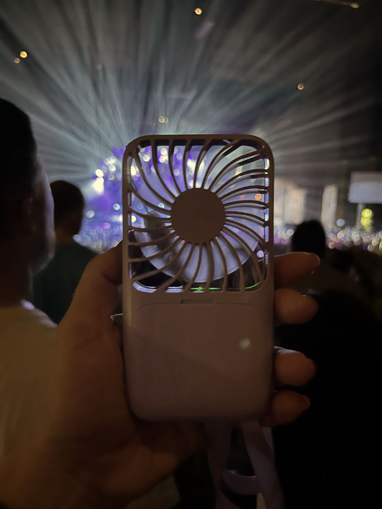 kim is holding her small purple portable fan at the concert. The stage lights behind it appear as if they are coming from the fan itself.