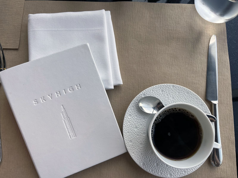 A top-down shot of the white skyhigh menu with a cup of coffee next to it. The coffee cup and saucer are white with a delicate pattern of different sized circles.