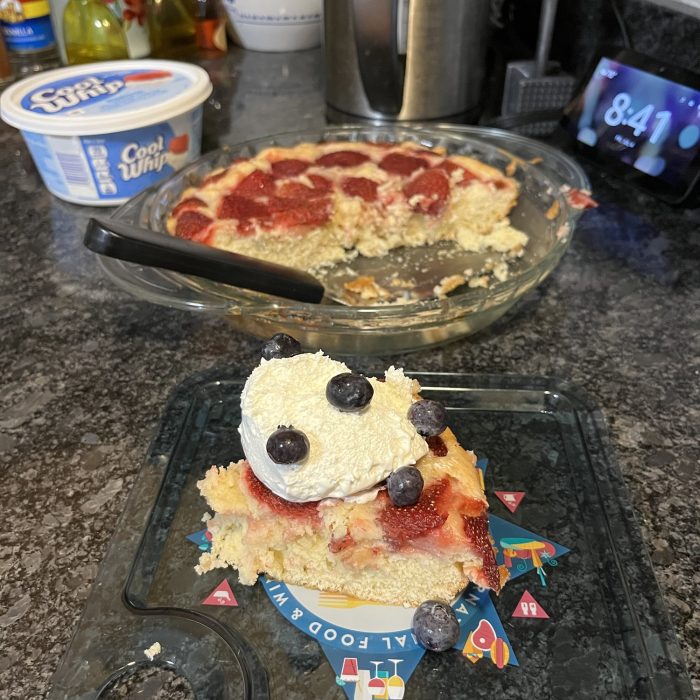 an unaesthetic photo of Smitten Kitchen's Strawberry cake with a dollop of Cool Whip and some blueberries on top. In the background is the entire cake and the tub of cool whip.