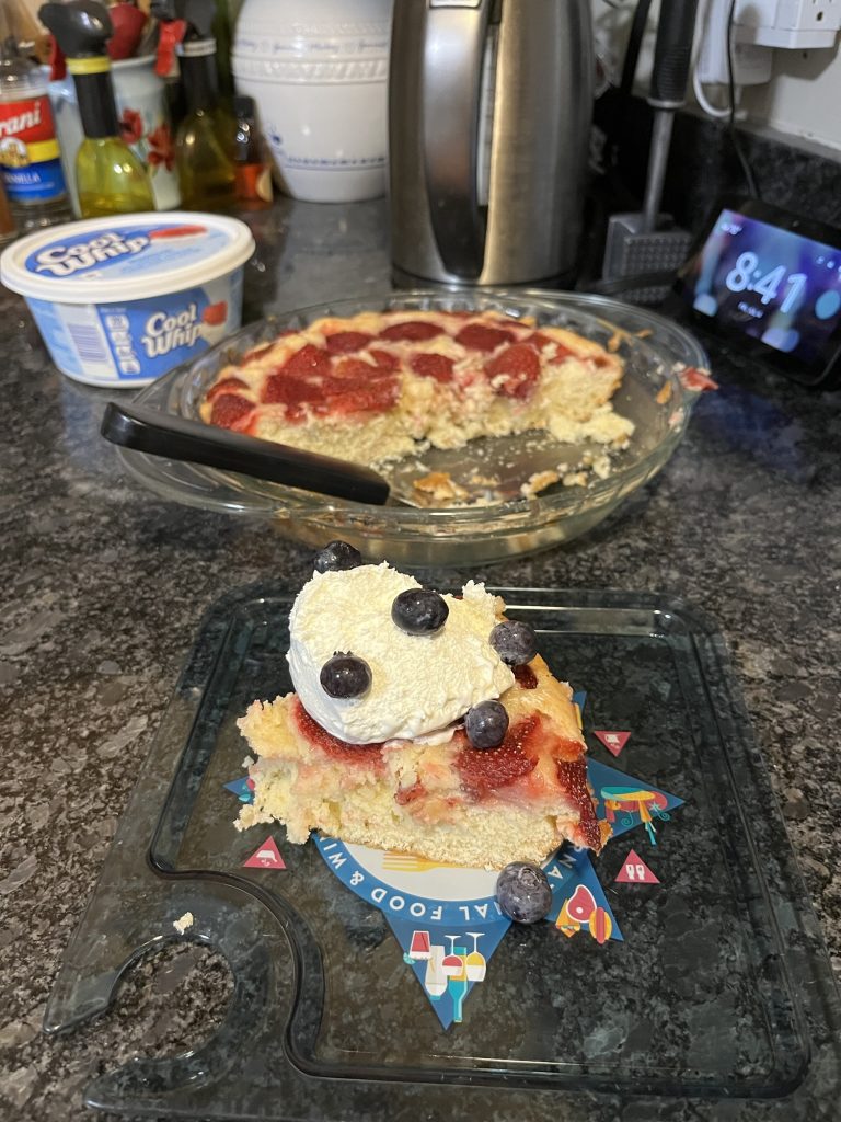 an unaesthetic photo of Smitten Kitchen's Strawberry cake with a dollop of Cool Whip and some blueberries on top. In the background is the entire cake and the tub of cool whip.