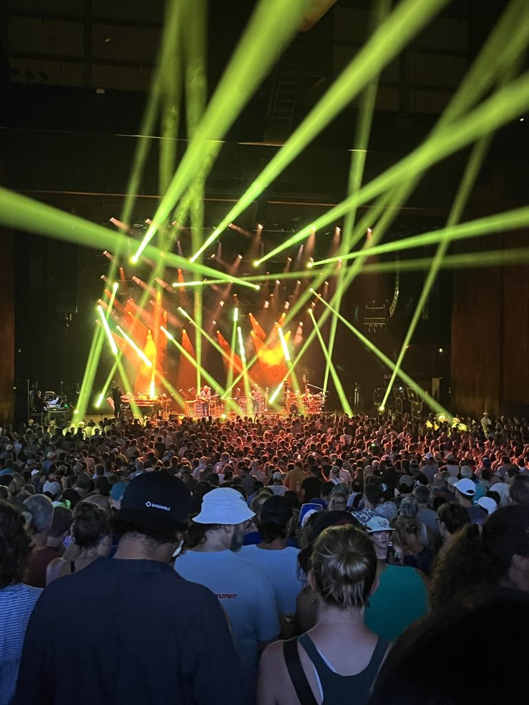The band Phish is onstage. There are many beams of greenish yellow light and a very happy crowd. Lots of smoke.