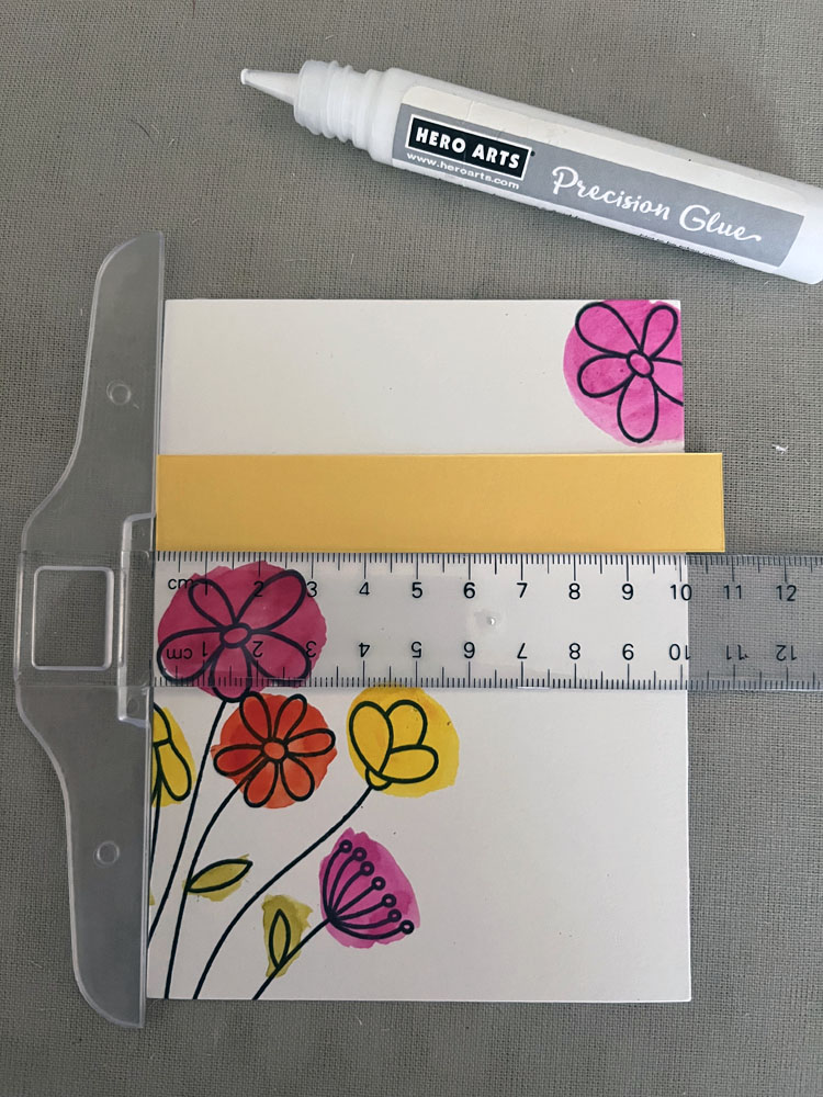 card in progress: stamped flowers with colorful spots of pink, yellow, and orange behind them, a yellow strip of paper about 1/4 down from the top  with a clear plastic t-square ruler helping to position it straight