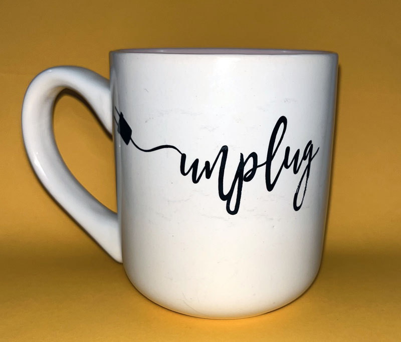 a white mug with the word unplug printed on it in a cursive script. the line that feeds into the "u" has a plug on it