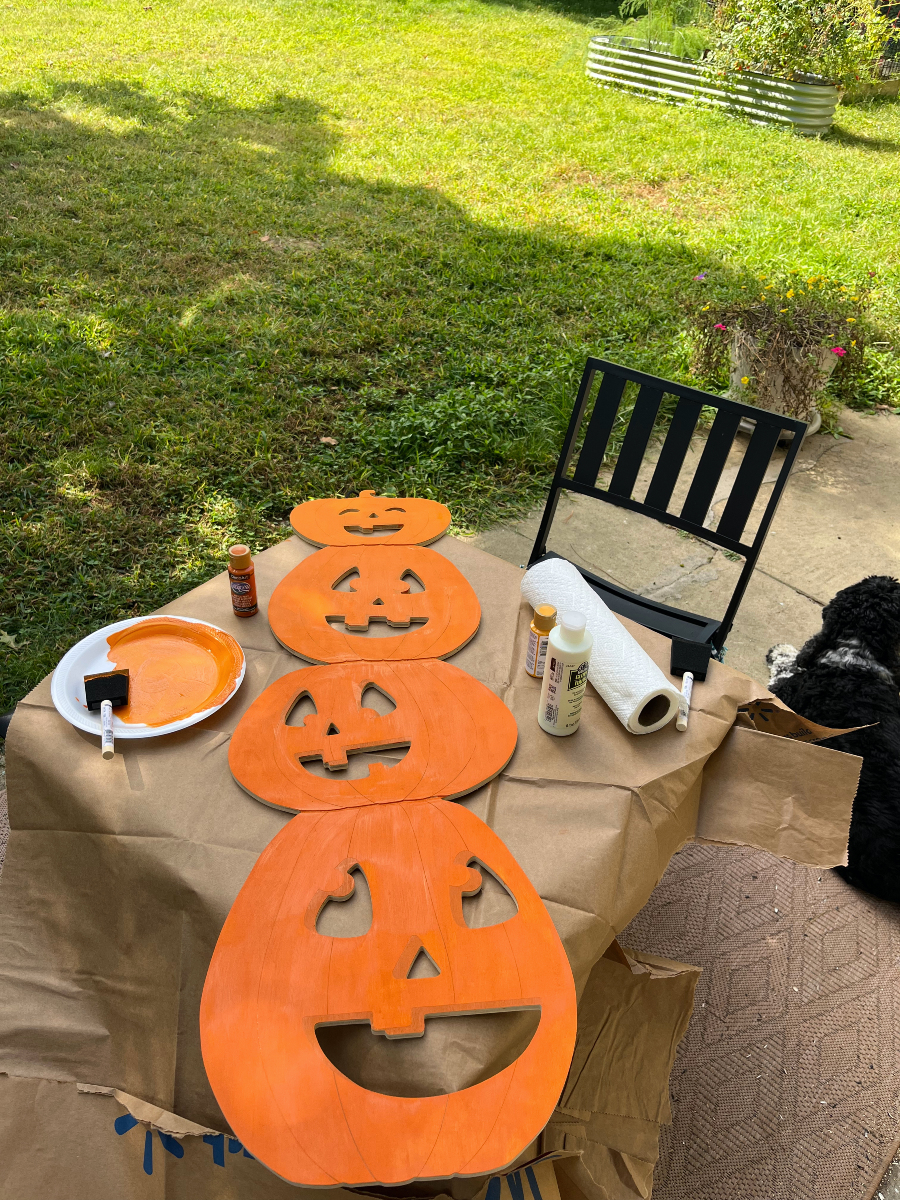 a flat stack of pumpkins made out of thin wood, they are painted orange and are lying flat on a table that's covered in brown paper. There is also a paper plate with orange paint on it