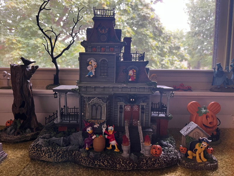 a Halloween village house on a ledge in front of a window. Huey, Louie, and Dewey are hanging out of the upstairs windows. Donald and Daisy are dressed up and outside below. Pluto has a pirate hat on.
