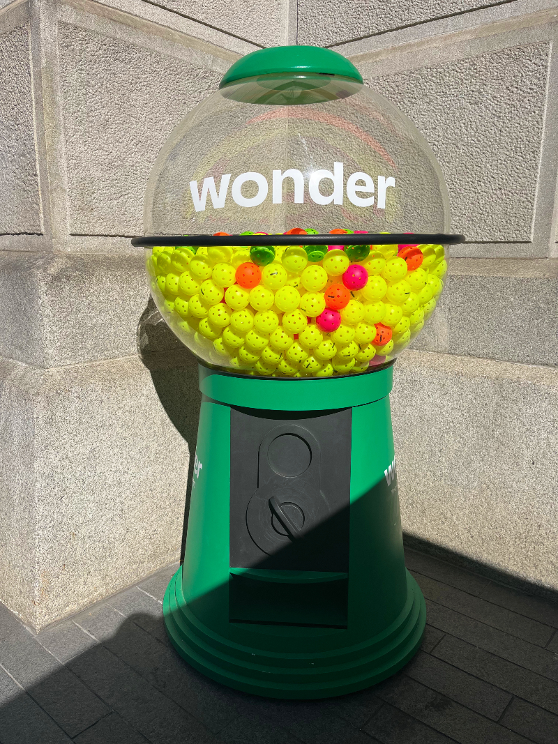 a giant gumball machine filled with mostly yellow and a few orange pickleballs.